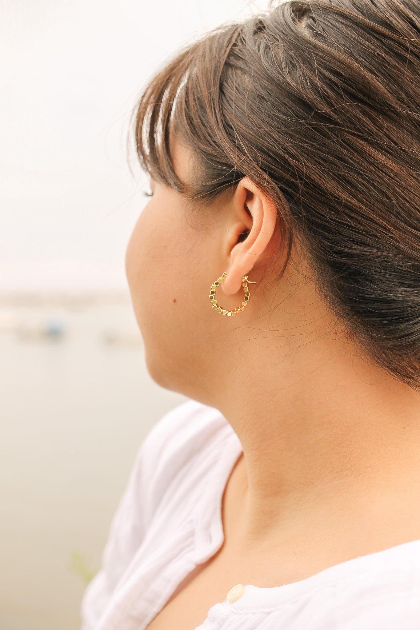 A woman with medium-length hair wears a gold hoop earring, gazing away from the viewer. She is by a body of water, creating a serene background.