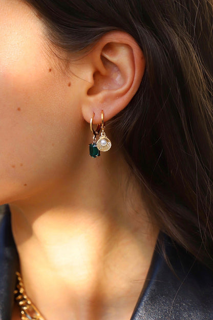 A close-up of an ear adorned with a gold hoop earring featuring two charms: a dark green gemstone and a pearl, set against a softly blurred background.