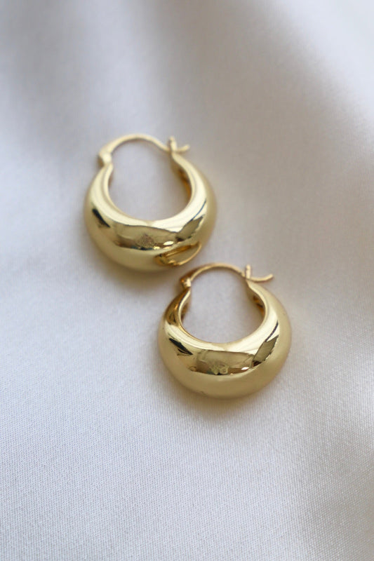 Two shiny gold hoop earrings rest on a soft, light-colored fabric. The earrings are smooth and round, with a crescent shape and a clasp hinge, showcasing their polished surface.