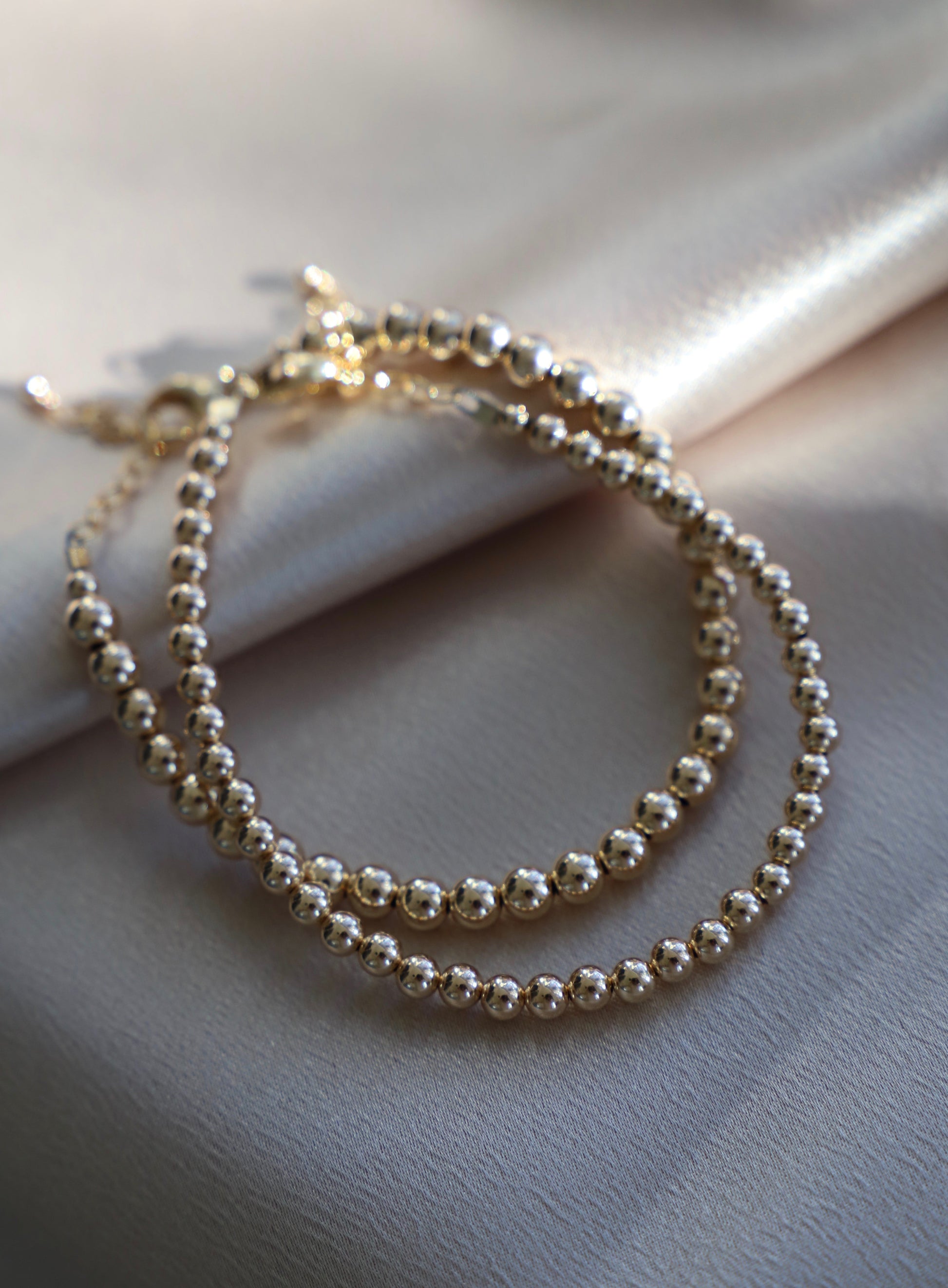 A gold beaded necklace is arranged elegantly on a soft, silky beige fabric, reflecting light and casting soft shadows, highlighting its smooth, polished surfaces.