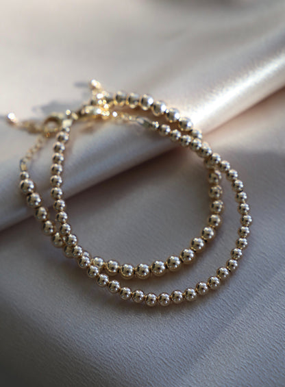 A gold beaded necklace is arranged elegantly on a soft, silky beige fabric, reflecting light and casting soft shadows, highlighting its smooth, polished surfaces.