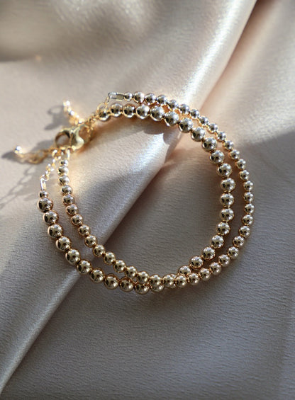 A shiny gold bracelet made of interlinked round beads is elegantly resting on a soft, beige satin fabric, reflecting light and creating a sense of luxury.