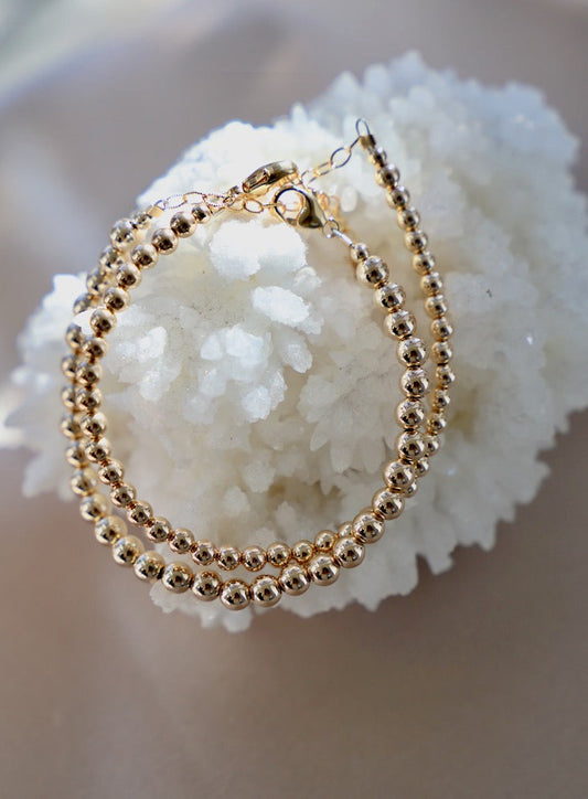 A circular gold bracelet, composed of small beads, rests on a textured white crystal. The sunlight reflects off the beads, enhancing their shine against a soft, neutral background.