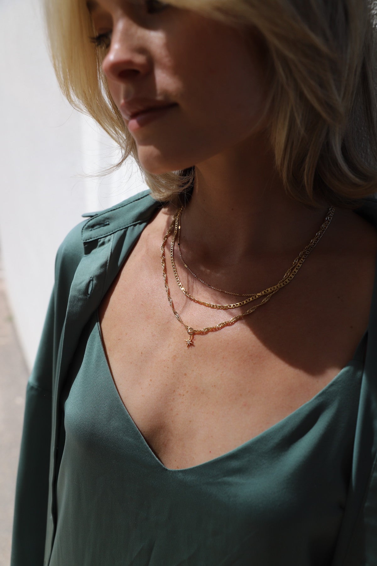 A woman with shoulder-length blonde hair wears layered gold necklaces. She is slightly angled, showcasing a green satin top and delicate jewelry, illuminated by soft sunlight against a light background.