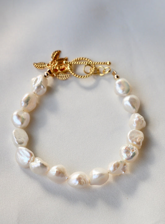 A pearl bracelet lies on a soft, light-colored fabric. The bracelet features irregularly shaped white pearls linked together, with a decorative gold clasp adorned with intricate details.