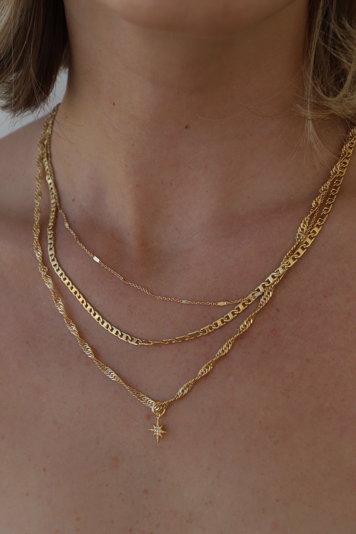 A set of layered gold necklaces drapes elegantly around a person's neck, showcasing various chain styles and a central star-shaped pendant, against a neutral skin tone background.