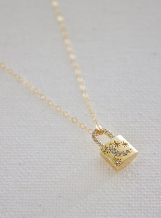 A delicate gold necklace features a small, shiny padlock pendant adorned with stars and tiny gems. It hangs against a soft, light-colored fabric background.