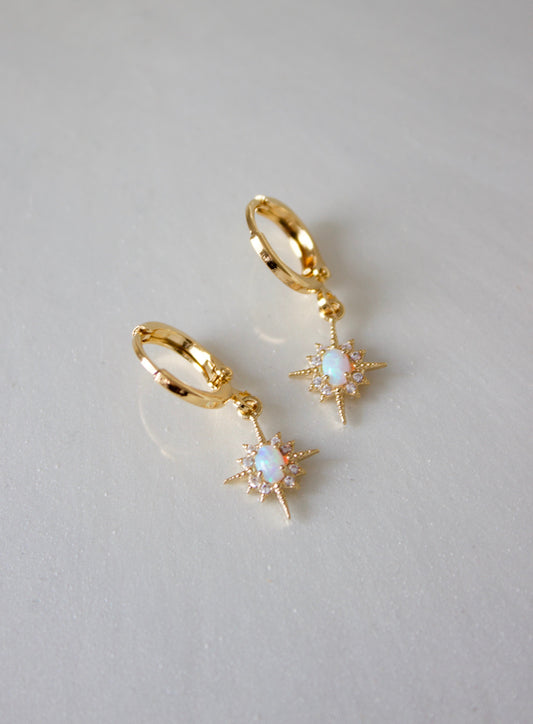 Gold hoop earrings with starburst designs hang from a smooth, light-colored surface, each featuring an opalescent stone at the center, reflecting light in a delicate display.