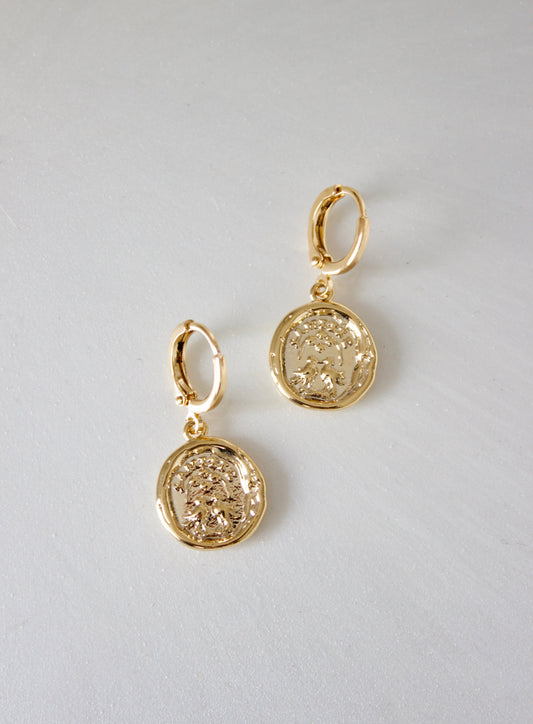 Gold earrings with circular pendants featuring intricate engravings hang against a smooth, light-colored surface, showcasing their reflective shine and detailed design.