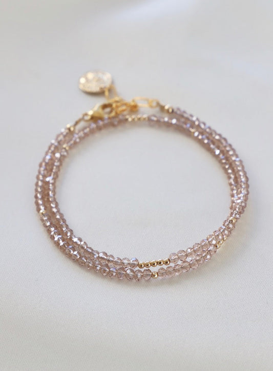 A delicate beaded bracelet made of shimmering light pink crystals is displayed, featuring a gold clasp and a circular charm. It rests on a soft, neutral fabric background.