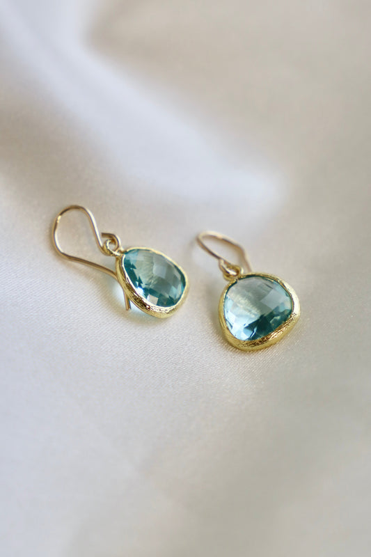 Two elegant earrings with teardrop-shaped, light blue stones are displayed against a soft, smooth white fabric background, showcasing their intricate gold settings and delicate hooks.