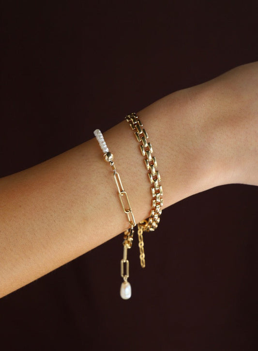 A wrist adorned with two gold bracelets and a white bead bracelet. The bracelets feature various links and a pearl drop, set against a dark brown background.