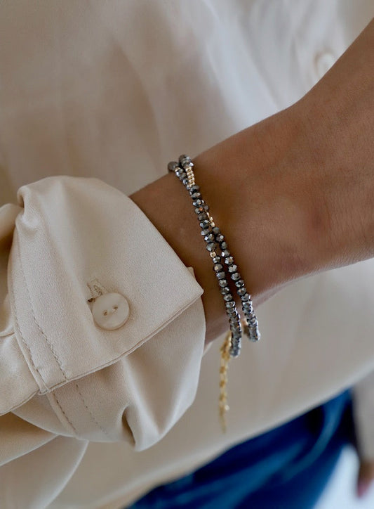 A hand with two beaded bracelets adorns the wrist, resting near a buttoned, light-colored shirt sleeve. The background is blurred, focusing attention on the jewelry and clothing.