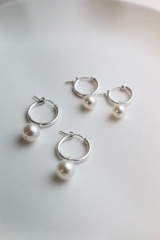 Hoop earrings with pearl drops rest on a smooth, light-colored surface. Their shiny metal contrasts nicely with the lustrous pearls, creating an elegant and modern accessory display.