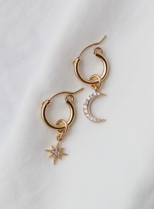 Two gold hoop earrings are displayed on a soft, light fabric. One earring features a hanging star design, while the other showcases a crescent moon adorned with sparkling stones.