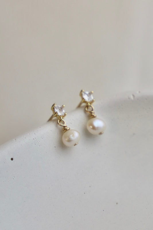 Two elegant earrings feature a clear gemstone atop a hanging pearl, resting on a light, textured surface. The soft background enhances the jewelry's delicate design.