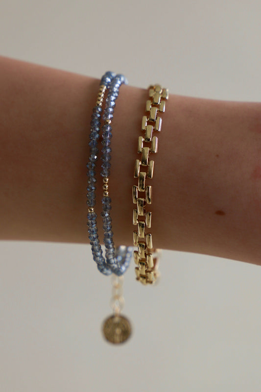 A wrist adorned with three bracelets: two delicate blue beaded strands and a shiny gold chain, against a soft, neutral background, showcasing the elegance of the accessories.