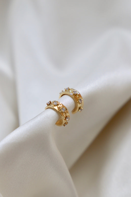 Two gold hoop earrings adorned with tiny decorative stones rest on a smooth, flowing piece of white fabric, reflecting light softly in a serene setting.