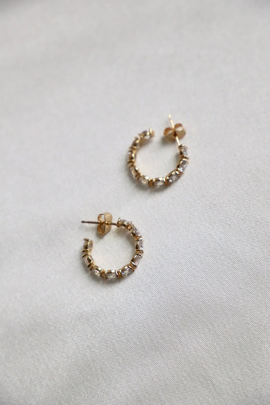 Two small gold hoop earrings adorned with rectangular gemstones rest on a smooth, light-colored fabric surface, showcasing their sparkling design and elegant style.