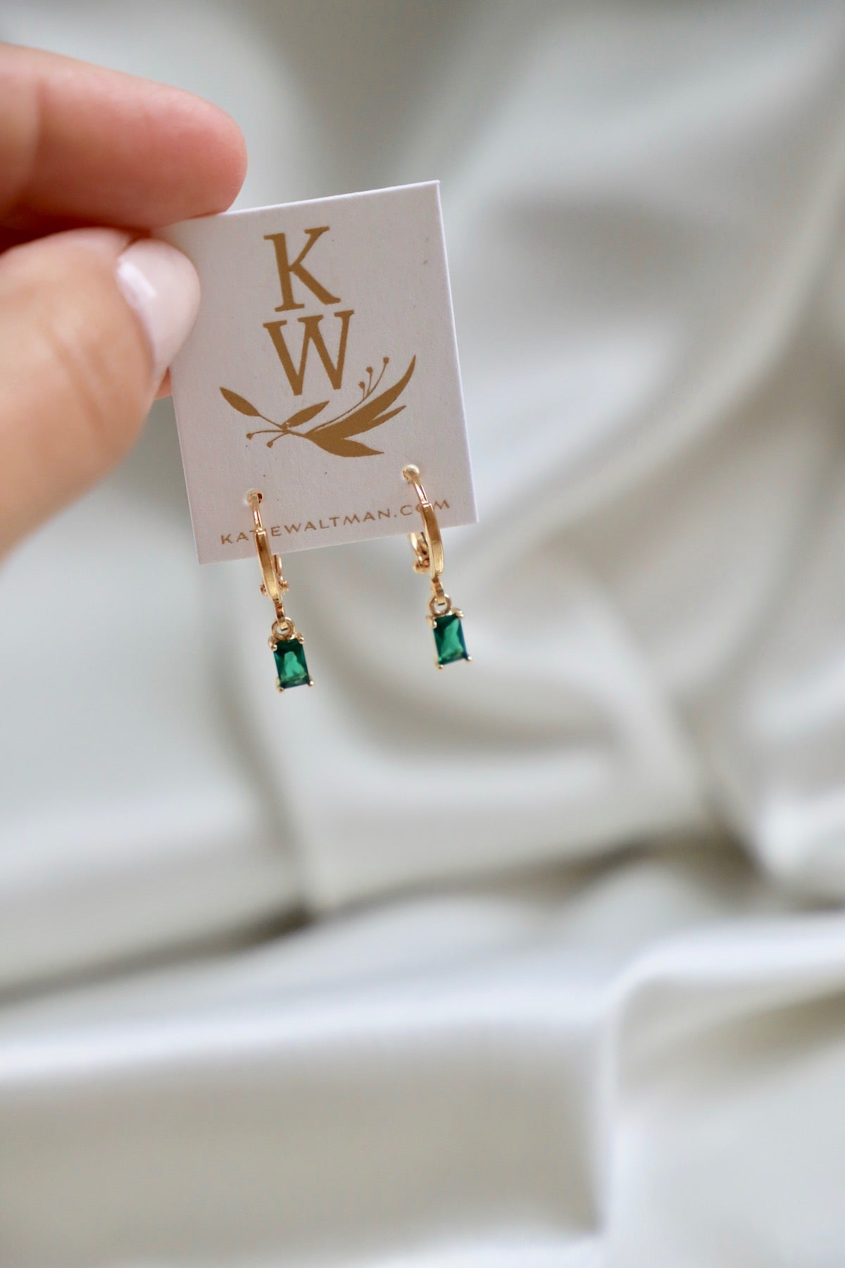 A pair of elegant gold earrings with green gemstones hang from a white display card featuring a logo. The background is soft and slightly wrinkled, suggesting a textile.