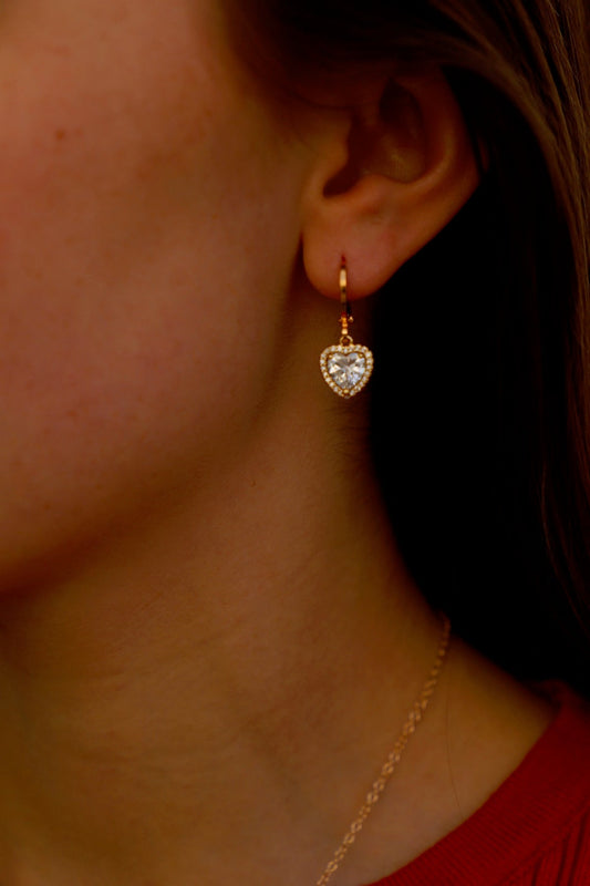 A heart-shaped earring, embellished with a central stone and surrounded by smaller gems, hangs from a woman's ear while she wears a red top, set against a blurred background.