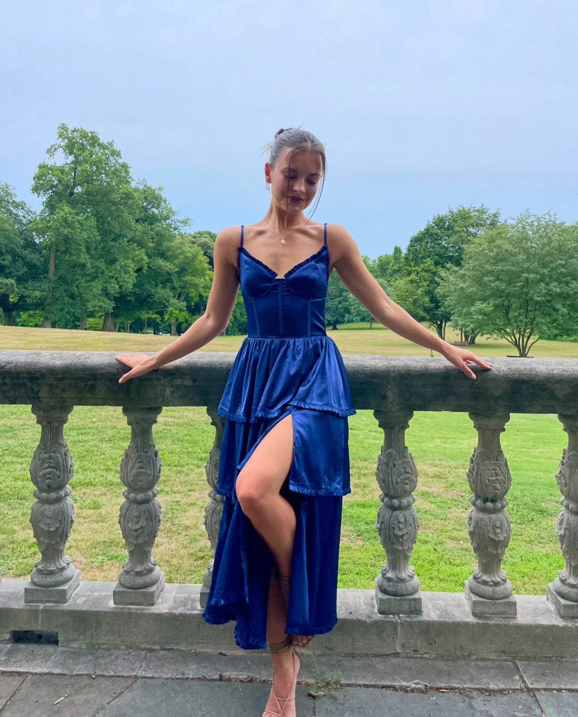 A woman wearing a blue satin dress stands by a stone railing, gazing downward. She is surrounded by a grassy landscape under a cloudy sky, creating a serene atmosphere.