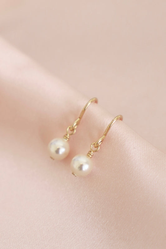 Two elegant gold hoop earrings, each adorned with a smooth, lustrous pearl, rest on a soft, pale pink satin background, highlighting their delicate design.