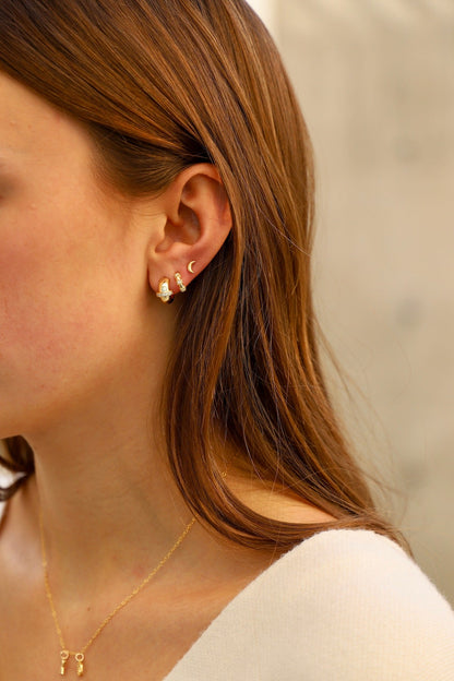 A young woman with long brown hair showcases multiple gold earrings in her ear. She wears a simple, light-colored top in a softly lit environment, emphasizing her jewelry.