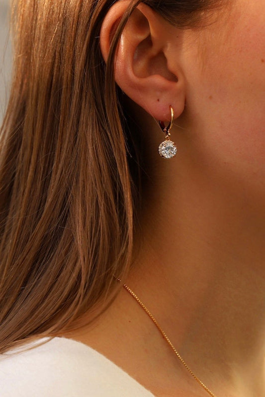 A stylish earring featuring a large, sparkling stone hangs from a woman’s ear, framed by long, flowing hair. She wears a delicate gold necklace, adding elegance to her look.