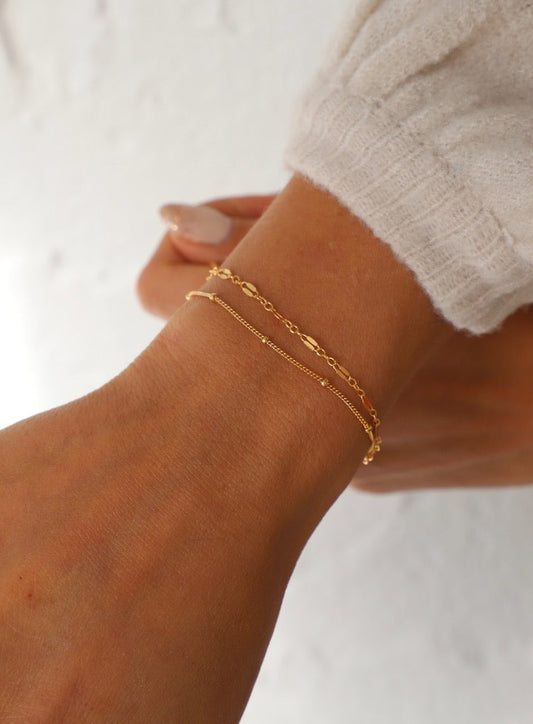 A slender wrist adorned with two delicate gold bracelets rests against a light-textured background, showcasing the accessories with a soft, natural light illuminating the scene.