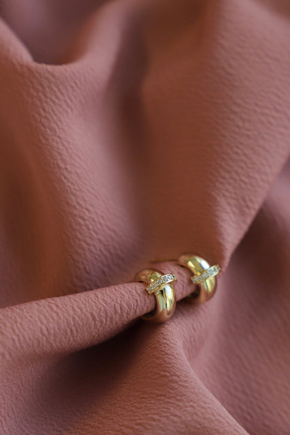 Two golden hoop earrings sit elegantly on a soft, rust-colored fabric. The earrings are adorned with small sparkling stones, contrasting beautifully against the textured background.