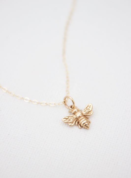 A delicate gold necklace features a bee pendant. The pendant is intricately designed with wings and a body, suspended from a fine chain, resting against a textured white background.