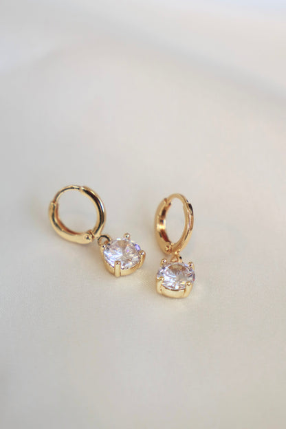 Two gold hoop earrings with clear, round gemstones hang gently on a soft, light-colored fabric, showcasing their elegant design and sparkling facets in a simple, refined setting.