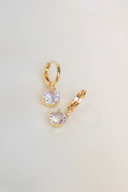 Two gold earrings with circular, sparkling gems are positioned on a smooth, light-colored surface, reflecting light and emphasizing their elegant design.