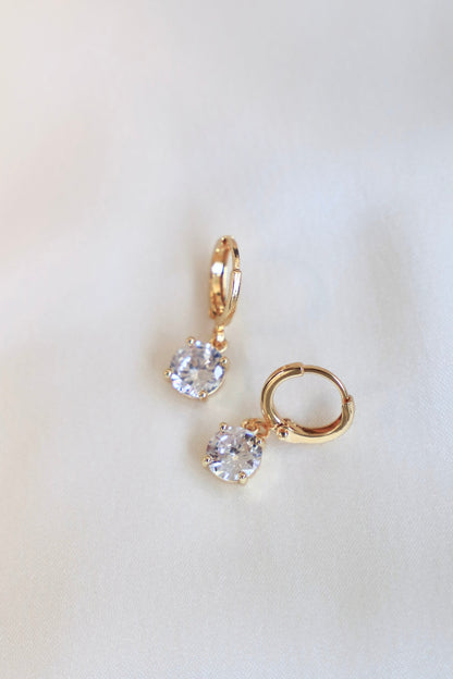 Two gold hoop earrings with clear gemstones hang on a soft, light-colored fabric, reflecting light and showcasing their sparkle. The design appears elegant and delicate.