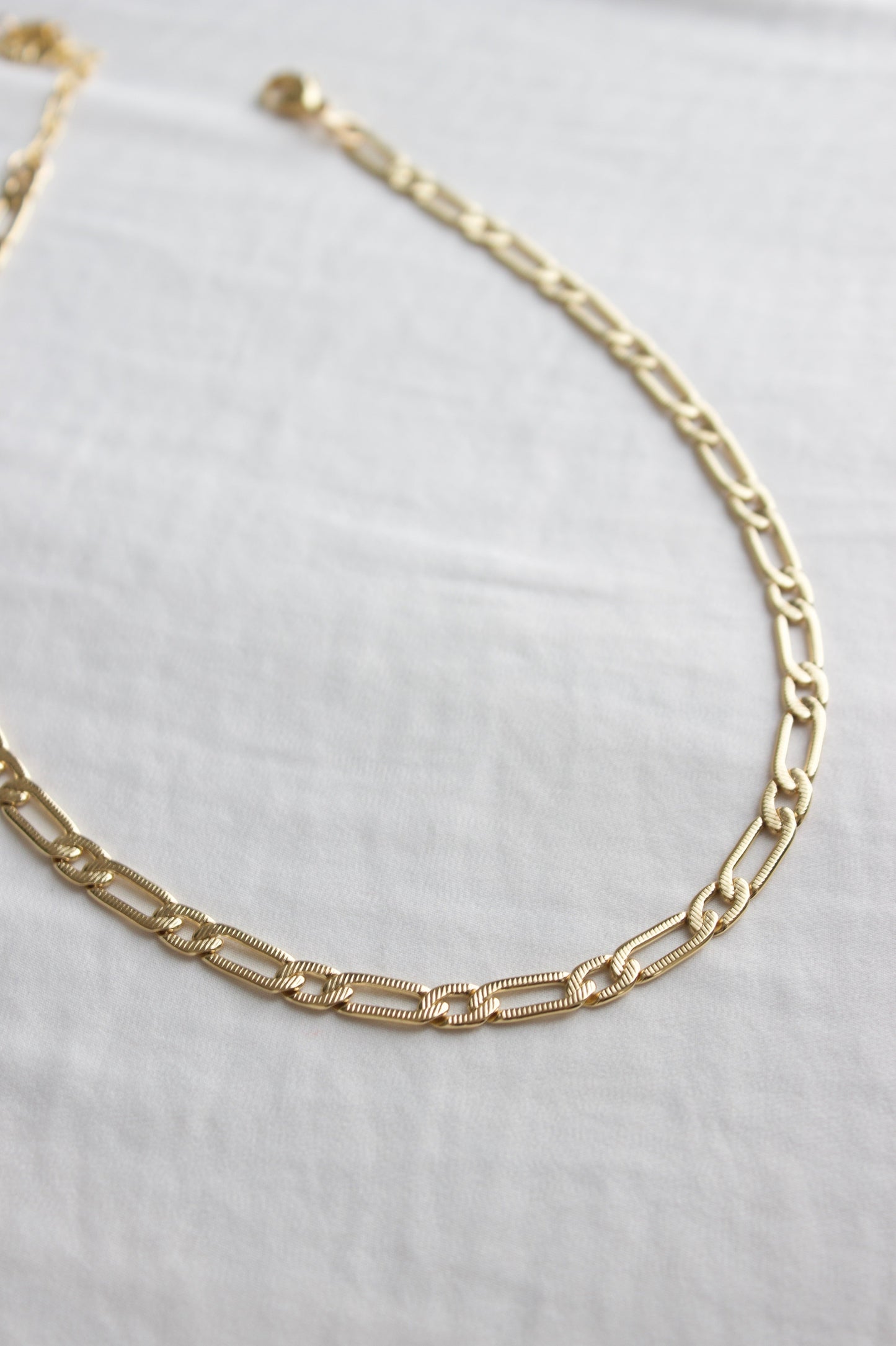 A gold chain necklace rests flat on a smooth, light-colored fabric. The links are alternating in design, contributing to an elegant appearance in a minimalistic setting.