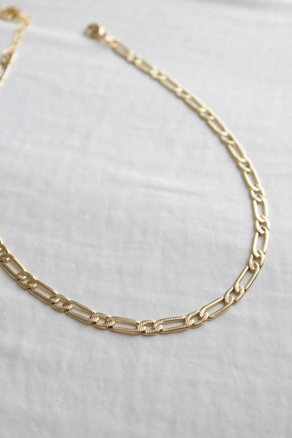 A gold chain necklace rests flat on a smooth, light-colored fabric. The links are alternating in design, contributing to an elegant appearance in a minimalistic setting.