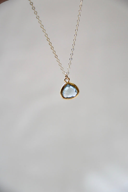 A delicate gold necklace with a teardrop-shaped blue gemstone hangs in the center, set against a soft, blurred background highlighting its elegance and simplicity.