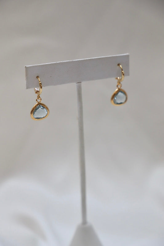 Gold-hued earrings with teardrop-shaped blue stones hang from a display stand. The background features a soft, neutral fabric, creating an elegant, simple presentation.