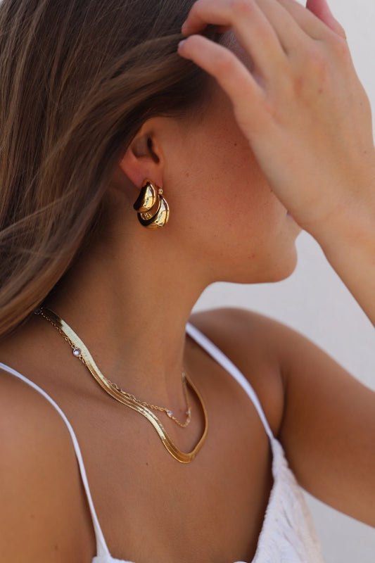 A woman with long hair brushes it aside, wearing stylish golden hoop earrings and layered necklaces. She is positioned against a plain light-colored background.