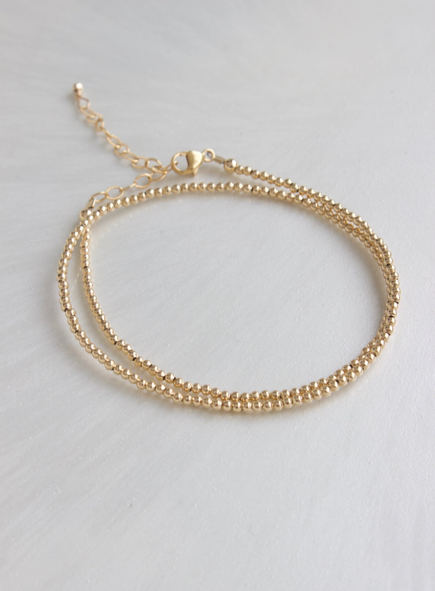 A delicate gold bracelet rests on a smooth, light surface, featuring two parallel strands of small spherical beads and a clasp for fastening.