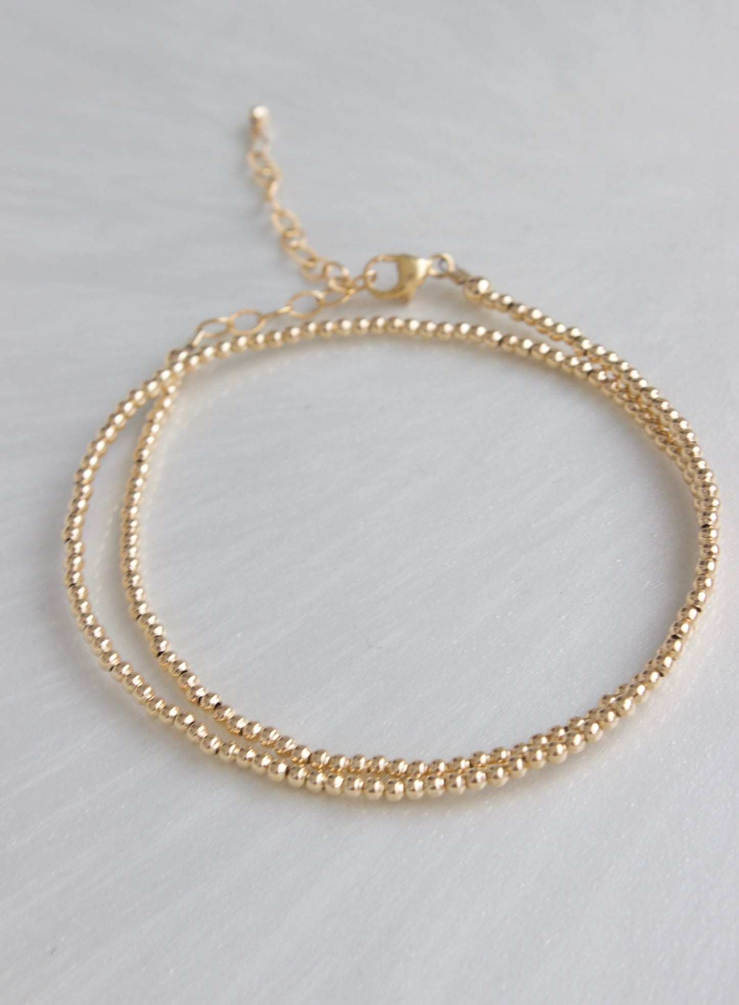 A delicate gold bracelet with two intertwined strands of small beads rests on a smooth, light-colored surface, showcasing its elegant design and craftsmanship.