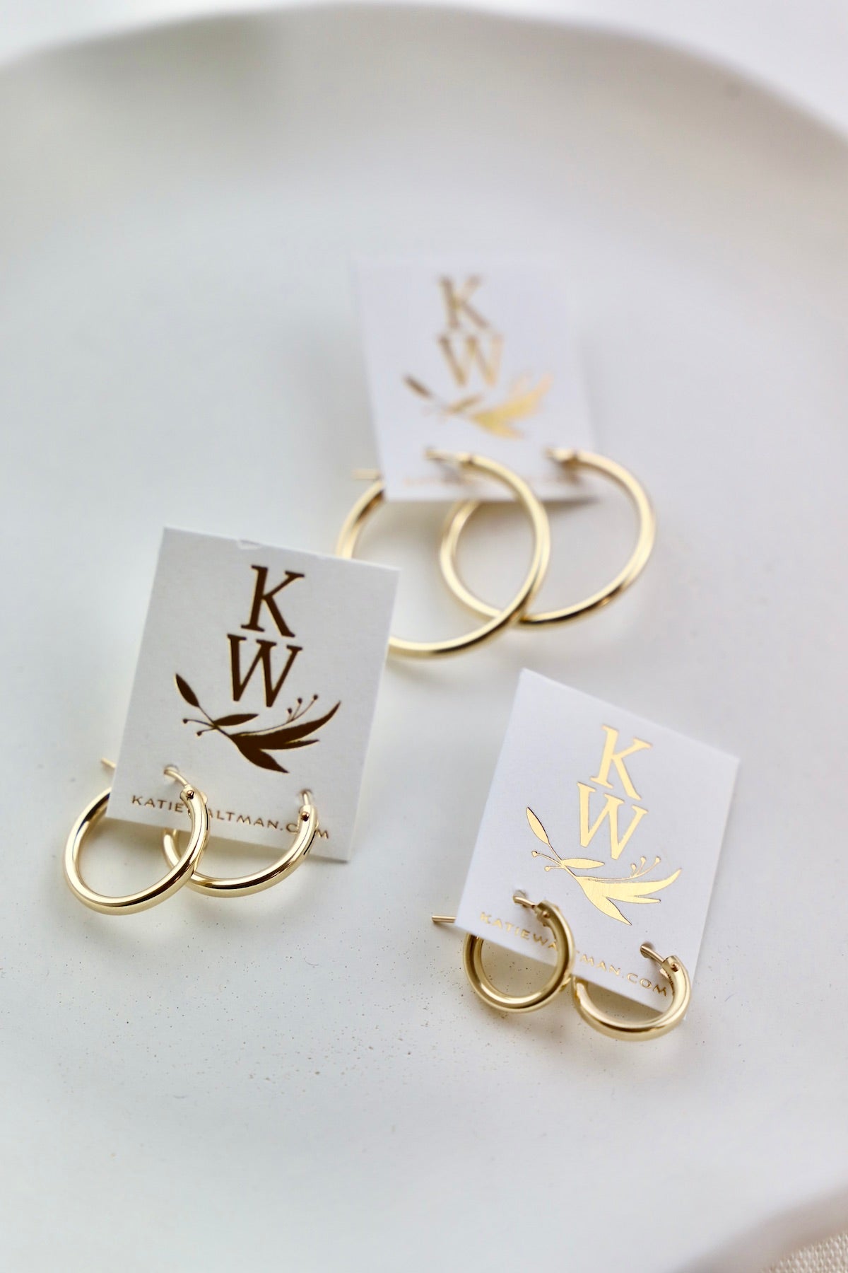 Three pairs of gold hoop earrings are displayed on white cards. Each card features a logo with "KW" and a decorative design, set against a soft, neutral background.