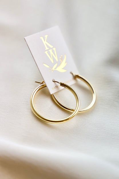 Gold hoop earrings are displayed on a light-colored card with a logo. The earrings rest on a soft, neutral fabric background, emphasizing their shine and design.