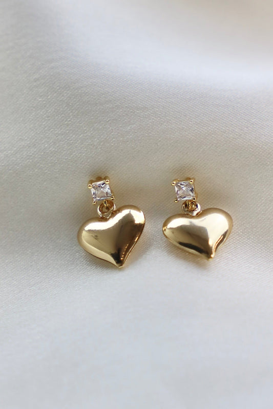 Two gold heart-shaped earrings with a small square clear stone above each heart rest on a smooth, light fabric background. The earrings have a shiny finish, reflecting the light.