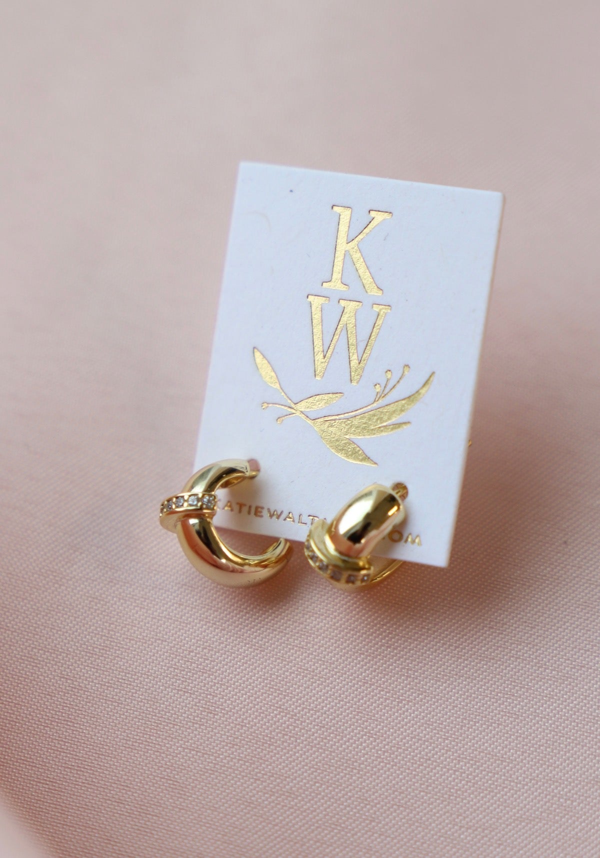 Gold hoop earrings with a small crystal detail are displayed on a white card featuring the initials "KW" and a decorative design, set against a soft pink background.