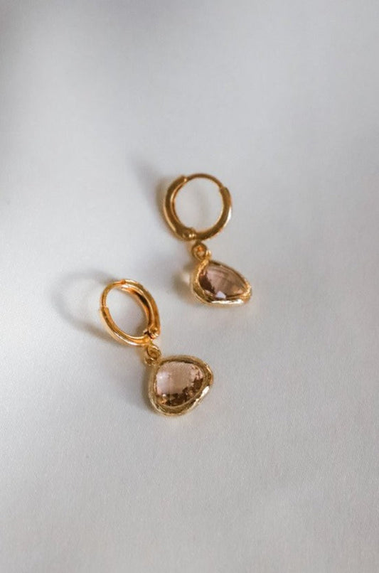 Gold hoop earrings feature a delicate drop design with teardrop-shaped, light pink gemstones. They rest on a soft, neutral background, emphasizing their elegant appearance.