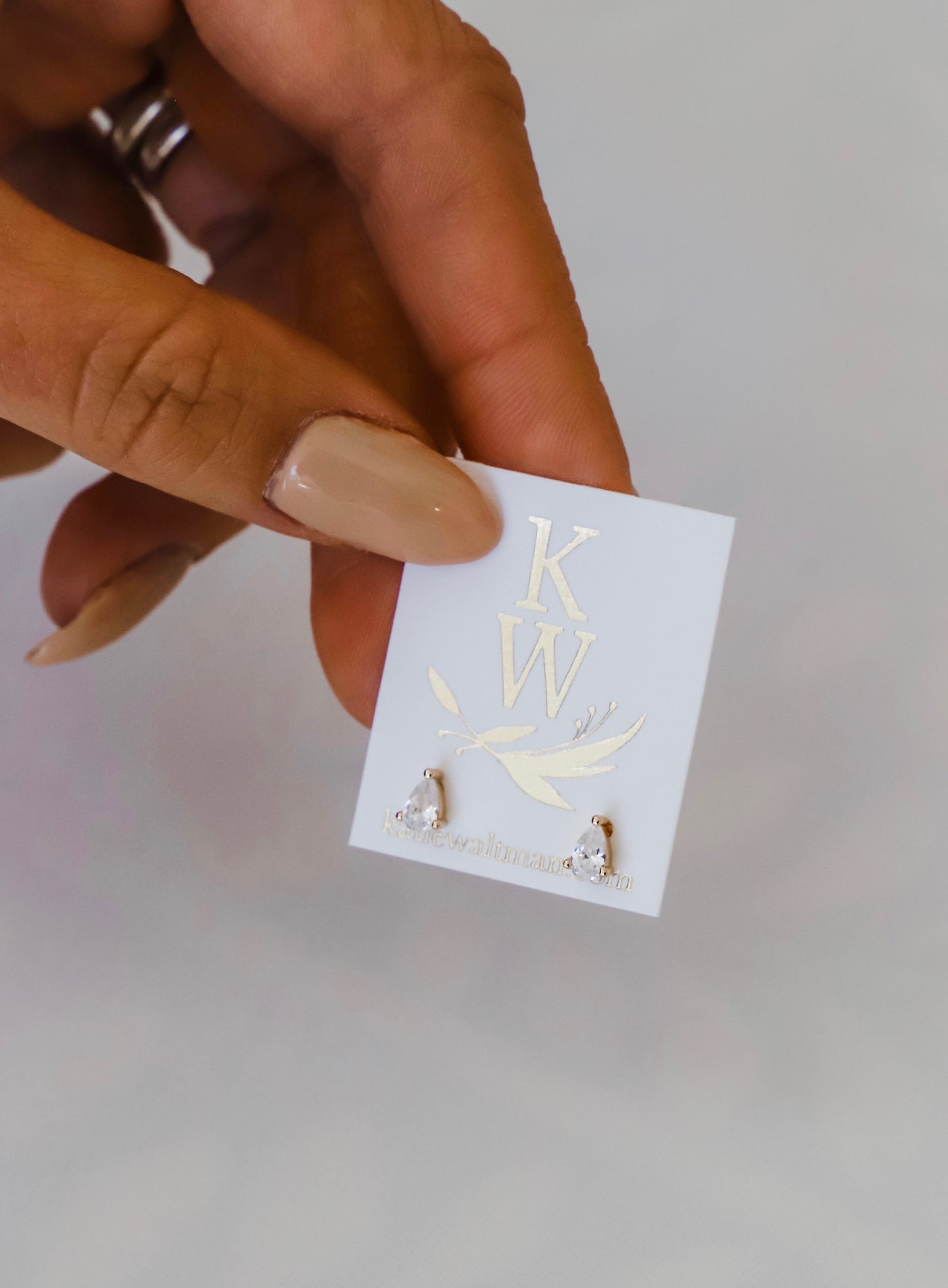 A person’s hand holds a card displaying two earrings. The earrings are teardrop-shaped and sparkly, while the card features the initials "KW" and a website URL, all set against a neutral background.