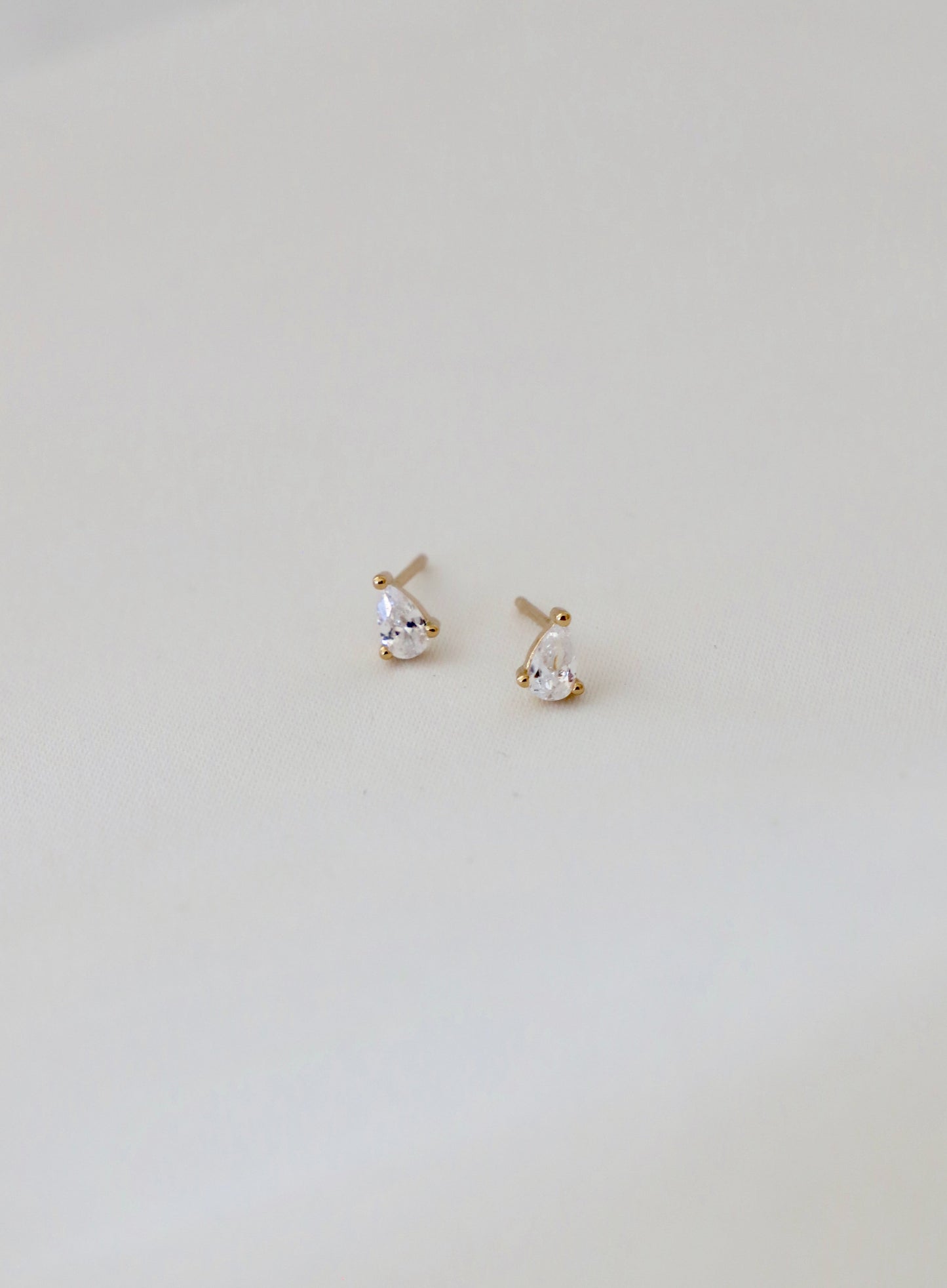 Two small, tear-shaped earrings with clear gemstones are displayed on a soft, light-colored fabric surface, showcasing their sparkling and elegant design.