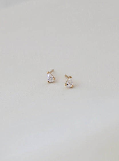 Two small, tear-shaped earrings with clear gemstones are displayed on a soft, light-colored fabric surface, showcasing their sparkling and elegant design.